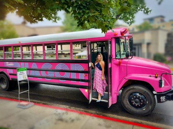 Bright pink vintage-style shuttle bus parked on a downtown curb under leafy trees, a smiling woman in a floral dress stepping out onto wet pavement.