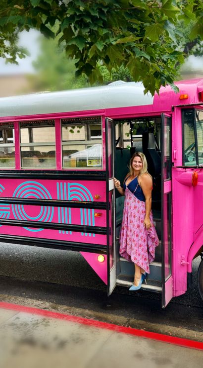 Bright pink vintage-style shuttle bus parked on a downtown curb under leafy trees, a smiling woman in a floral dress stepping out onto wet pavement.