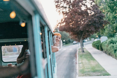Smiling person in an orange headscarf leaning out of a turquoise vintage van on a quiet suburban residential street at dusk, string lights inside and friends with drinks visible through the window.
