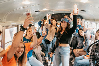 Group of young adults smiling and raising canned drinks inside a lively converted bus during a road-trip celebration.