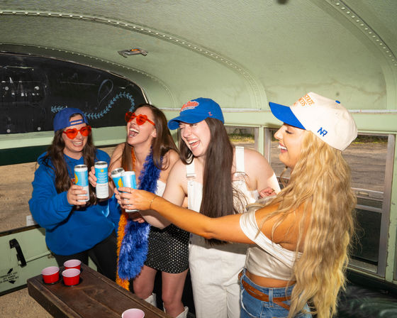 Four friends laughing and toasting canned drinks inside a converted green school bus at an outdoor party, wearing caps, heart-shaped sunglasses, a feather boa and casual summer outfits.