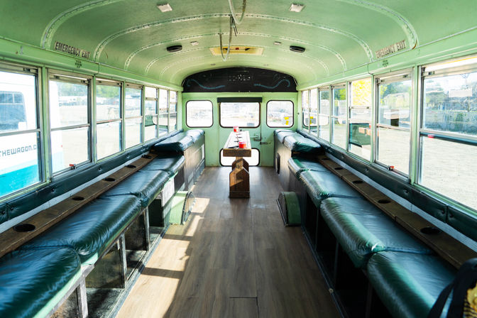 Sunlit retro converted school bus interior with curved green ceiling, long green vinyl bench seats along both sides, wood-plank floor, center aisle and small wooden table with red cups, large windows.