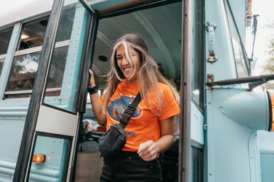 Beaming young woman stepping out of a light-blue converted school bus, wearing an orange team t‑shirt and a black crossbody bag.
