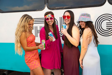 Four young women in colorful summer outfits laughing and holding canned drinks beside a teal-striped retro tour bus