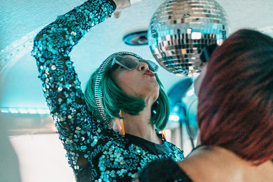 Person in a green bob wig and sequined teal dress reaches up toward a disco ball under blue neon lights, wearing oversized sunglasses and orange earrings with a red-haired friend blurred in the foreground.
