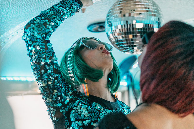 Person in a green bob wig and sequined teal dress reaches up toward a disco ball under blue neon lights, wearing oversized sunglasses and orange earrings with a red-haired friend blurred in the foreground.