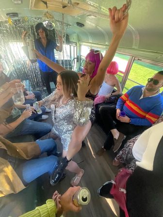 Party bus interior with a group of adults dancing under a disco ball and metallic streamers, colorful wigs and sequined outfits, people holding drinks and sunlight through the windows.