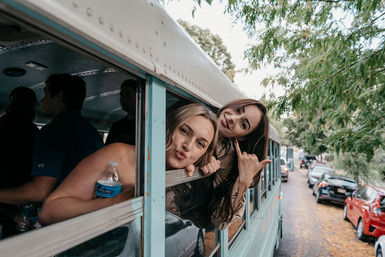 Two friends playfully leaning out of a light blue bus window, smiling and posing on a tree-lined street with parked cars.