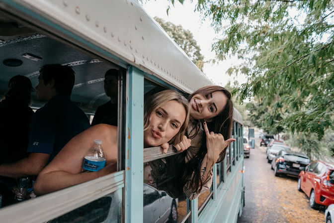 Two friends playfully leaning out of a light blue bus window, smiling and posing on a tree-lined street with parked cars.