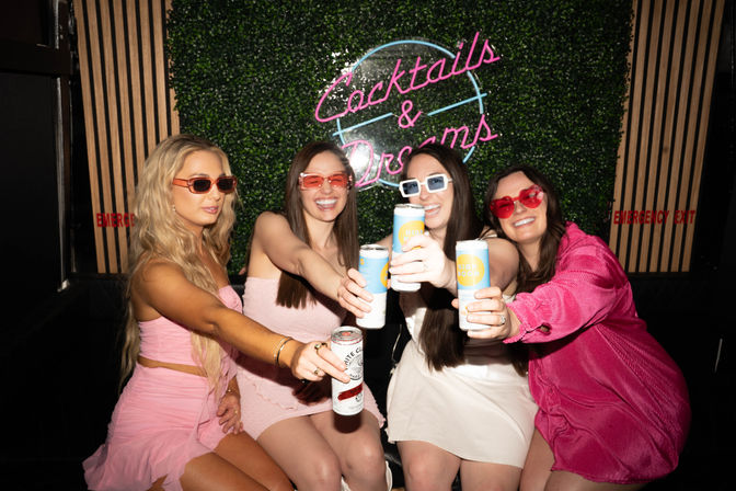 Four women toasting canned drinks in a bar lounge photo booth under a neon "Cocktails & Dreams" sign — pink outfits and playful sunglasses.