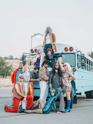 Playful group of adults in colorful 1970s-inspired costumes and wigs posing on and around a light blue bus in an outdoor parking lot, featuring sequins, bell-bottoms and retro boots.
