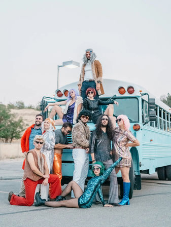 Playful group of adults in colorful 1970s-inspired costumes and wigs posing on and around a light blue bus in an outdoor parking lot, featuring sequins, bell-bottoms and retro boots.