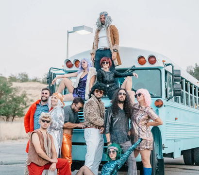 Playful group of adults in colorful 1970s-inspired costumes and wigs posing on and around a light blue bus in an outdoor parking lot, featuring sequins, bell-bottoms and retro boots.