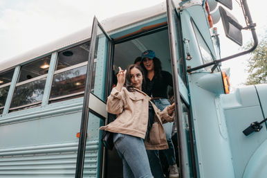 Two young women stepping off a light-blue converted school bus, one flashing a peace sign in a tan corduroy jacket and jeans for a casual road-trip vibe.