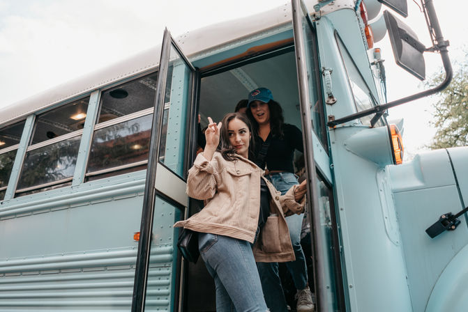 Two young women stepping off a light-blue converted school bus, one flashing a peace sign in a tan corduroy jacket and jeans for a casual road-trip vibe.