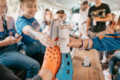 Group of friends on a party bus clinking canned drinks and bumping orange and blue slip-on clogs over a wooden bench