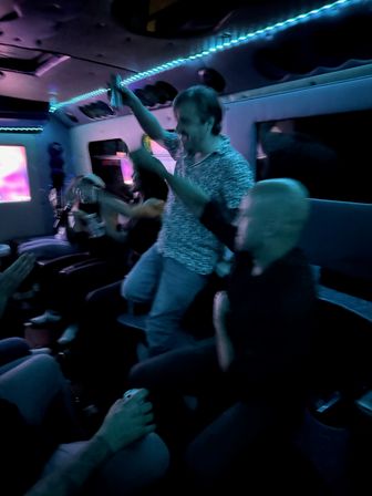 Group of people partying inside a neon-lit party bus, cheering and dancing with drinks under blue LED strip lights