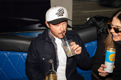 Two friends in a blue quilted bar booth on a night out — man in a white cap sipping a cocktail from a plastic cup while holding a bottle, woman in sunglasses laughing and holding a canned drink.