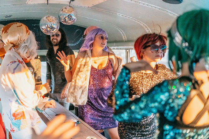 Glam party on a decorated bus: people in colorful wigs and sequin outfits dancing under disco balls