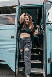 Laughing person in a black crop top and ripped jeans stepping out of a turquoise vintage bus on a street, showing a dragonfly belly tattoo — playful road-trip vibe.