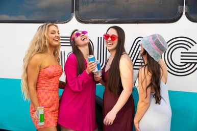 Four young women laughing and holding canned drinks in front of a colorful party bus, wearing bright summer dresses, sunglasses and a striped bucket hat — outdoor summer festival or road-trip vibe.