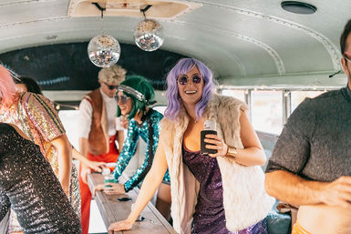People in colorful costumes partying inside a retro bus with hanging disco balls; a smiling person in a purple wig, round sunglasses and faux-fur vest holds a canned drink.