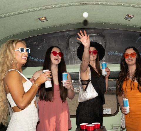 Four women in summer dresses and heart-shaped sunglasses play beer pong inside a retro party bus, tossing a ping-pong ball over red solo cups while holding canned drinks.