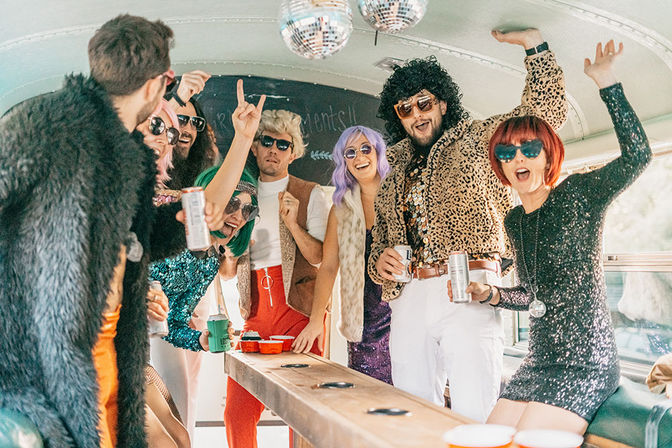 Group of friends in retro 1970s disco outfits and colorful wigs partying inside a decorated party bus with drinks and disco balls