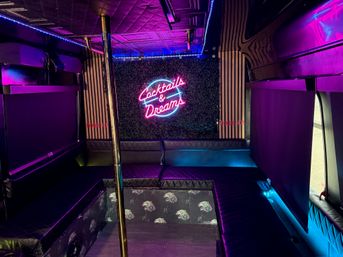 Vibrant neon-lit party bus interior with quilted black leather bench seating, brass dance pole, LED accent lights, and a "Cocktails & Dreams" neon sign.