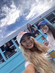 Group of women tailgating beside a light-blue bus, smiling with canned drinks and wearing Boise State Broncos caps under a bright cloudy sky