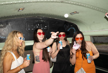 Four women in colorful sunglasses holding canned drinks and a photo prop inside a retro green party bus interior, playful road-trip vibe