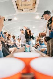Crowded party-bus interior with young adults playing beer-pong-style game, tossing a ball toward red cups on a long table while others hold cans and cheer.