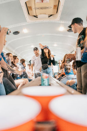 Crowded party-bus interior with young adults playing beer-pong-style game, tossing a ball toward red cups on a long table while others hold cans and cheer.
