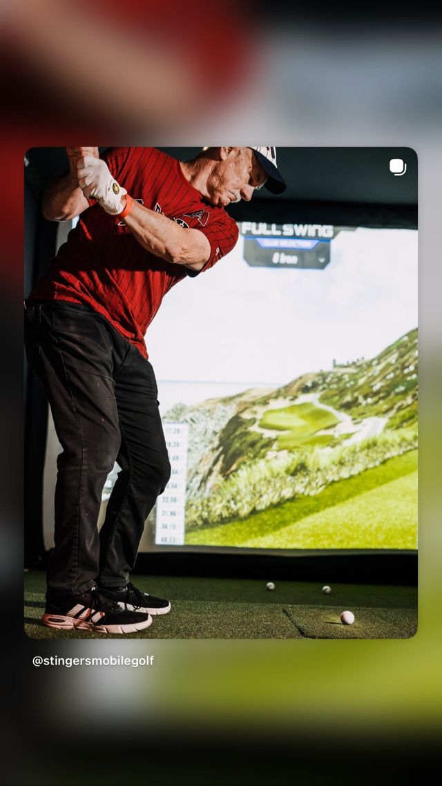 Golfer in red shirt mid-swing at an indoor golf simulator, aiming at a virtual coastal links hole on the screen with golf balls on the turf