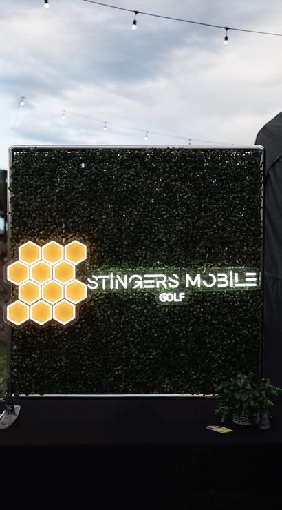 Honeycomb LED cluster and glowing mobile-golf sign on a boxwood backdrop at an outdoor evening festival, with hanging string lights and a cloudy sky overhead.