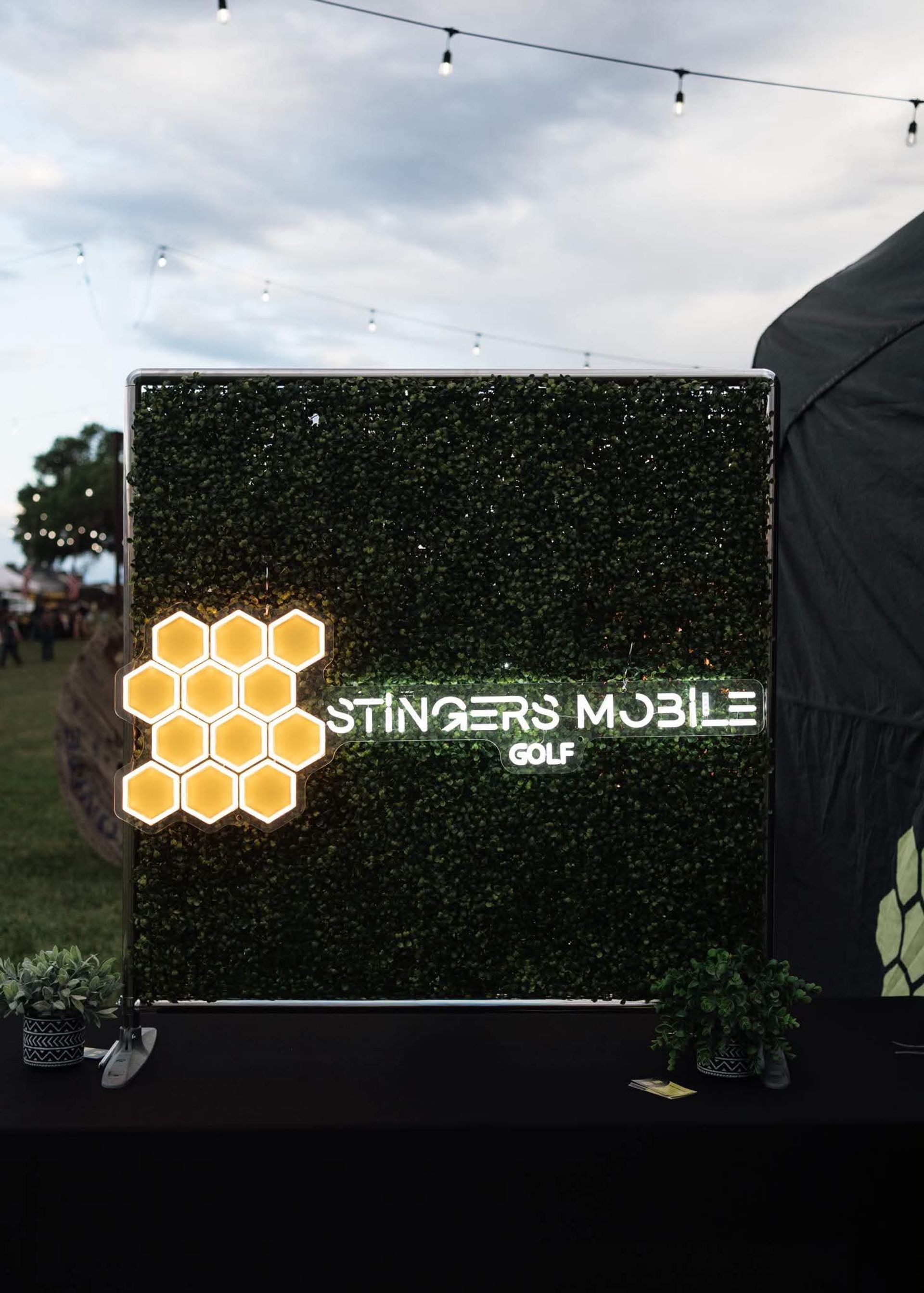 Honeycomb LED cluster and glowing mobile-golf sign on a boxwood backdrop at an outdoor evening festival, with hanging string lights and a cloudy sky overhead.