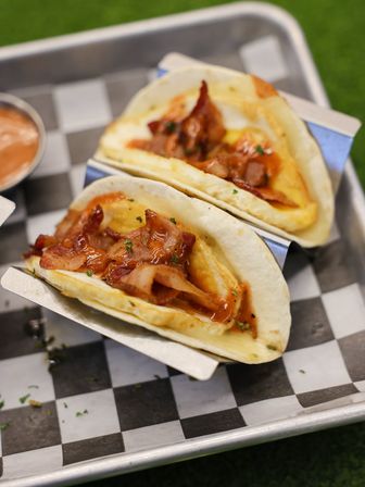 Two bacon-and-egg breakfast tacos with melted egg and crispy bacon drizzled in sauce inside soft flour tortillas on a metal tray lined with black-and-white check paper — brunch comfort food close-up.