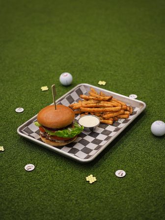 Burger with lettuce, tomato and red onion and seasoned fries with dipping sauce on a checkered tray, set on artificial putting-green turf with golf balls and tokens — playful golf-themed casual dining scene.