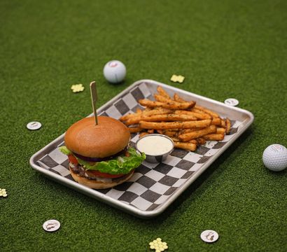 Burger with lettuce, tomato and red onion and seasoned fries with dipping sauce on a checkered tray, set on artificial putting-green turf with golf balls and tokens — playful golf-themed casual dining scene.