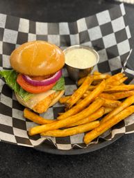 Crispy fish sandwich with lettuce, tomato and red onion on a shiny brioche bun, served with golden seasoned fries and a cup of tartar sauce on black-and-white checkered paper.