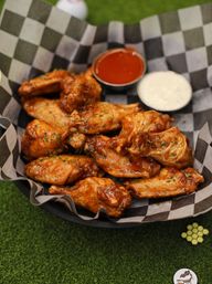 Crispy saucy buffalo chicken wings in a checkered-paper basket with hot sauce and ranch dip on green turf — a game-day snack