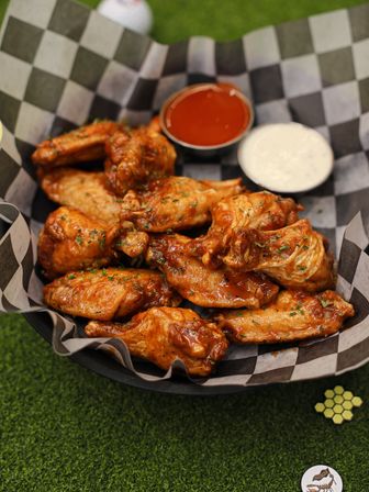 Crispy saucy buffalo chicken wings in a checkered-paper basket with hot sauce and ranch dip on green turf — a game-day snack