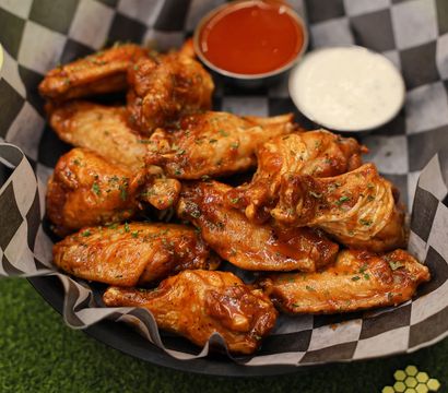 Crispy saucy buffalo chicken wings in a checkered-paper basket with hot sauce and ranch dip on green turf — a game-day snack