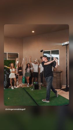 Friends cheering as a man swings a golf club at a portable indoor golf simulator on artificial turf in a covered patio — lively home golf party scene