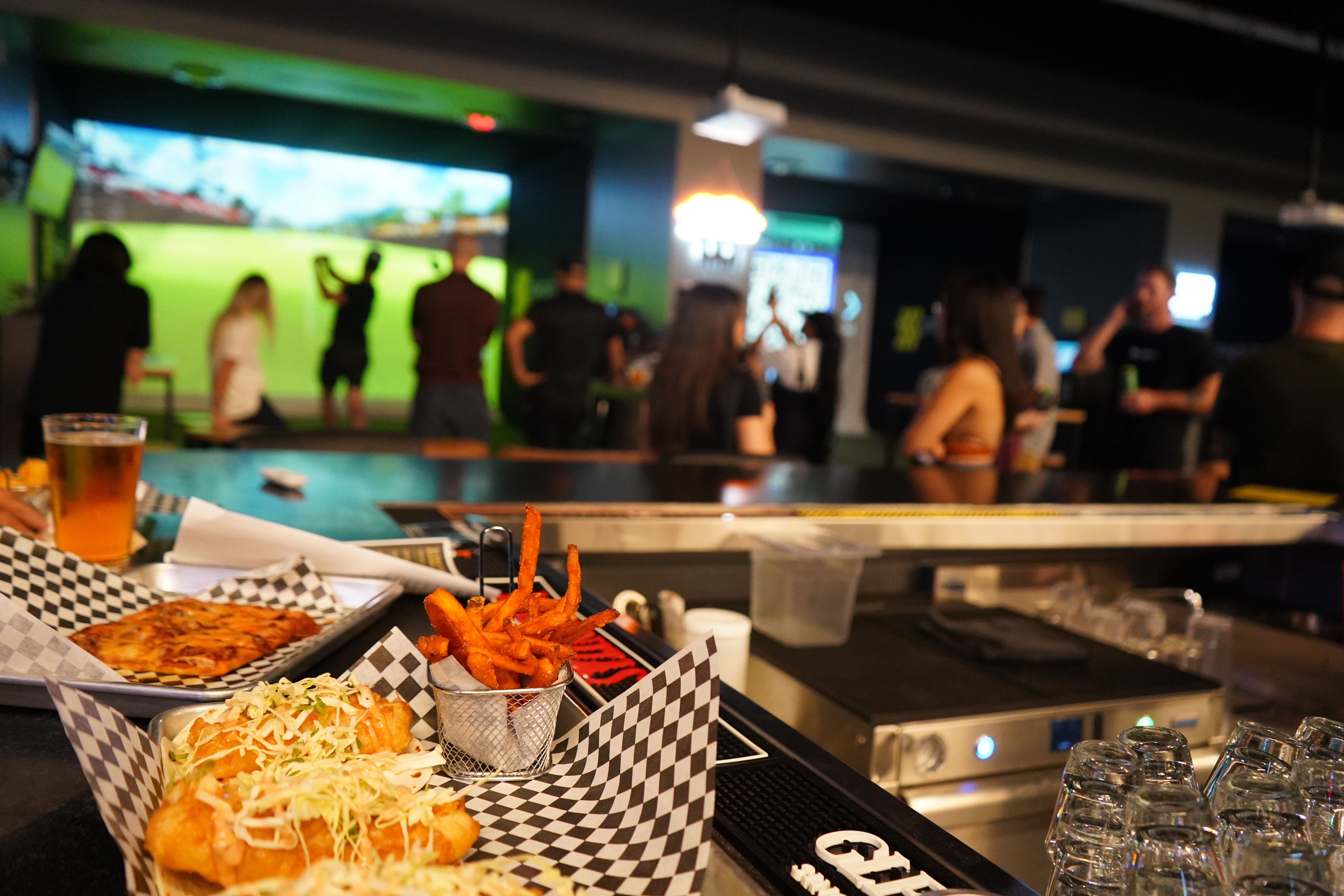 Bar counter close-up of fish tacos, sweet potato fries, pizza and beer at a lively indoor sports-simulator lounge with people playing virtual golf on a large screen