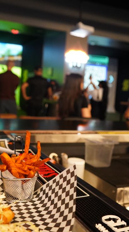 Bar counter close-up of fish tacos, sweet potato fries, pizza and beer at a lively indoor sports-simulator lounge with people playing virtual golf on a large screen