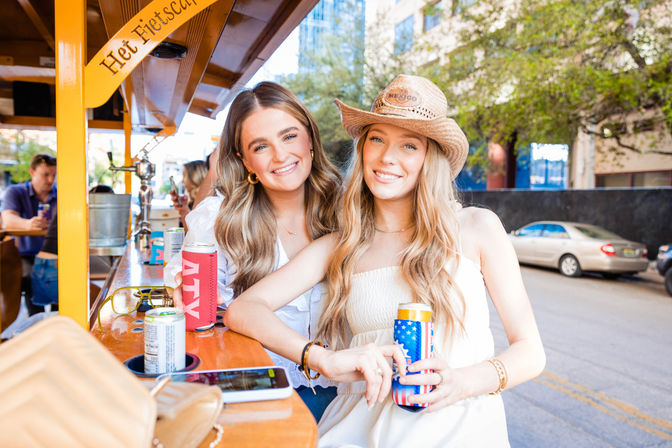 Two smiling friends in summer outfits, one wearing a straw cowboy hat, enjoying canned drinks with colorful koozies at a sunny outdoor bar counter on a downtown city street.