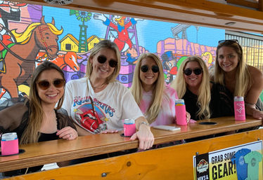 Five friends in sunglasses smiling at a wooden outdoor bar, each holding a canned drink in a pink koozie, with a bright cartoon-style street mural of horses and buildings behind them.