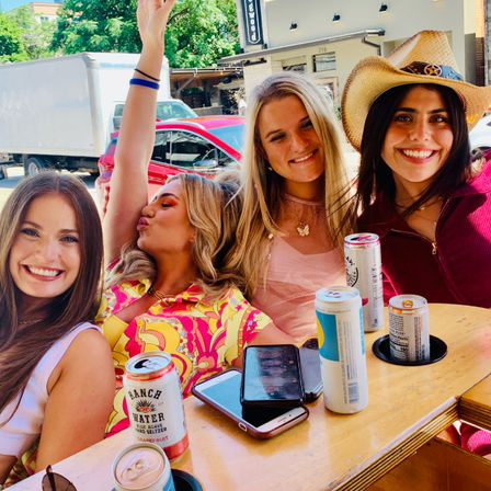 Four friends smiling and posing at a sunny outdoor patio table — one wearing a cowboy hat and another in a bright patterned dress, assorted canned seltzers and phones on the wooden table, parked cars and storefronts in the background.