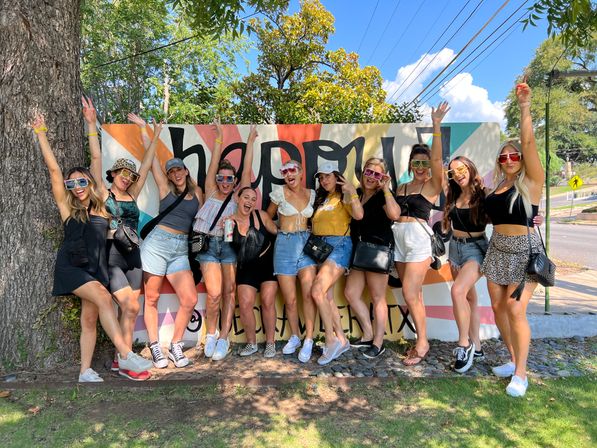 Group of friends in summer outfits and colorful sunglasses, arms raised and laughing while posing in front of a vibrant painted mural on a sunny tree-lined street.
